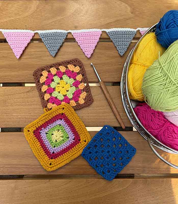 Gehäkelte Topflappen und Wimpelkette. Häkelnadel liegt daneben und Wollekneule in verschiedenen Farben sind zu sehen.